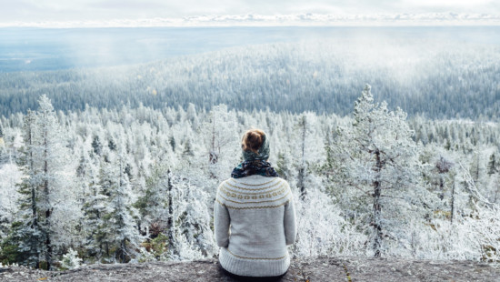 Nainen istuu kalliolla villapaita päällään. Taustalla näkyy metsää, joka on valkoisen huurteen peitoissa. Metsän yllä leijuu sumua. Kuva: Panu Mölsä / Metsähallitus