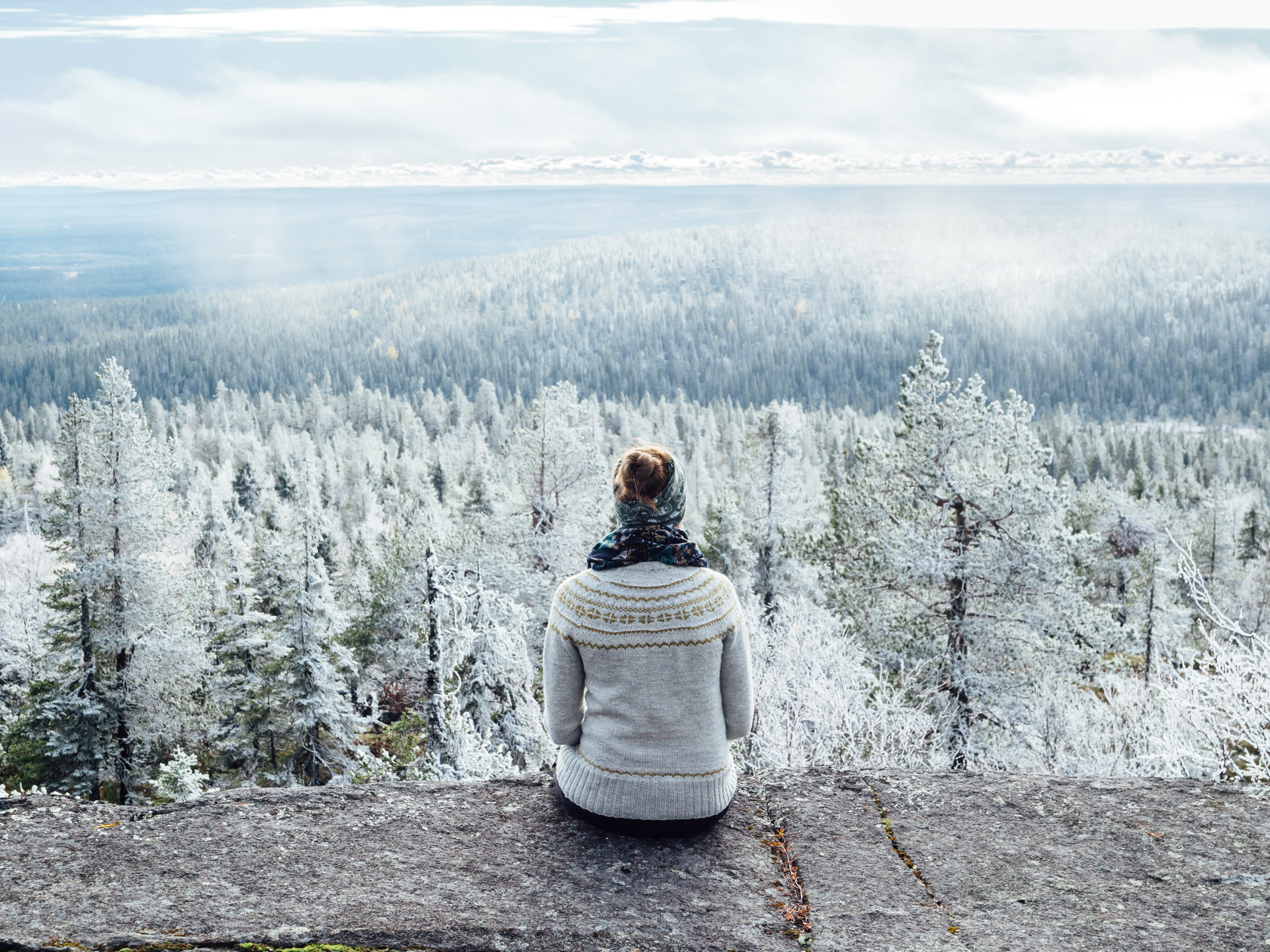 Nainen istuu kalliolla villapaita päällään. Taustalla näkyy metsää, joka on valkoisen huurteen peitoissa. Metsän yllä leijuu sumua. Kuva: Panu Mölsä / Metsähallitus